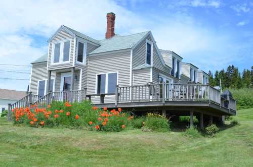 A large, light gray house with a deck, surrounded by vibrant orange flowers and green grass under a blue sky.