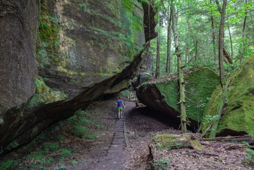 A hiker walks along a narrow trail surrounded by towering moss-covered rock formations in a lush green forest.