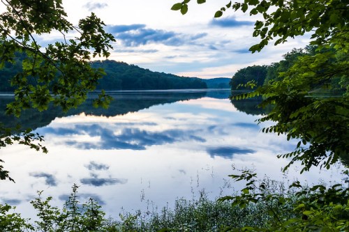 A serene lake reflects clouds and trees, framed by lush greenery under a soft, blue sky.