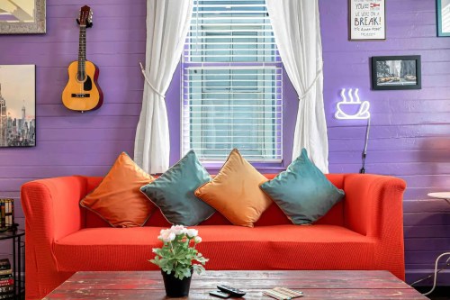 A cozy living room with a bright orange couch, colorful cushions, a guitar on the wall, and a coffee-themed neon sign.