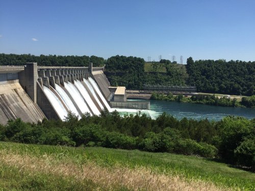 A dam releasing water into a river, surrounded by green trees and a clear blue sky.