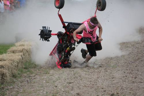 A person in a pink shirt falls off a modified lawnmower, kicking up dust and debris during a race.