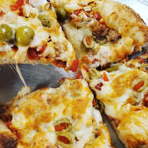 Close-up of a pizza with melted cheese, green olives, sausage, and diced red peppers, with a slice partially removed.