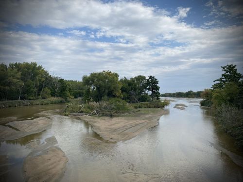 A serene river scene with sandy banks, lush trees, and a cloudy sky reflecting on the water's surface.