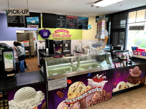 A colorful ice cream shop counter with various flavors, a menu board, and a staff member preparing orders.