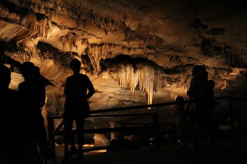 Silhouettes of people exploring a dimly lit cave with hanging stalactites and rock formations.