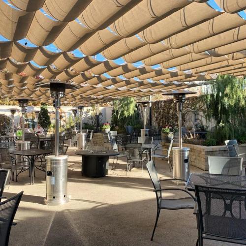 Outdoor dining area with shaded seating, heaters, and greenery under a blue sky.