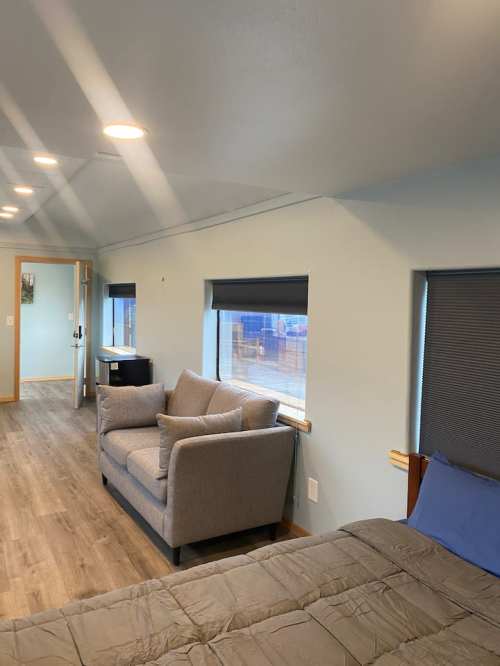 A cozy room featuring a gray sofa, a bed, and large windows with blinds, illuminated by overhead lights.