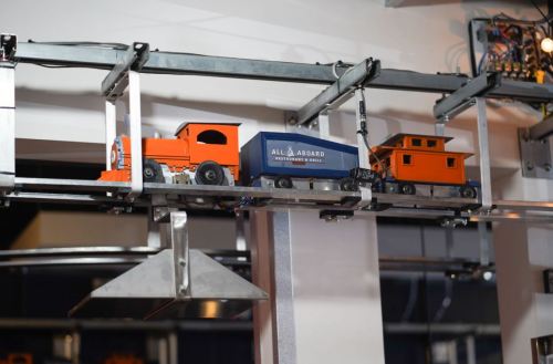 A toy train on a track, featuring an orange engine and blue cargo car, suspended above a restaurant setting.