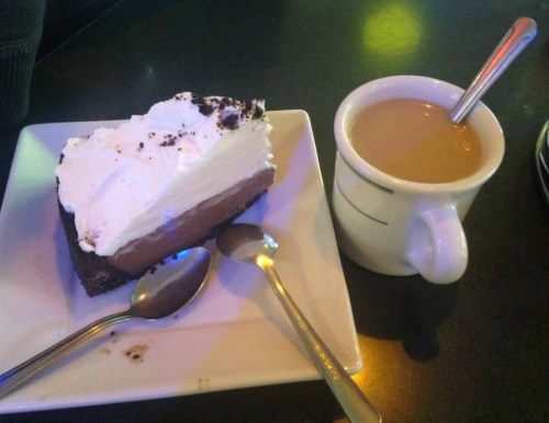 A slice of chocolate cream pie topped with whipped cream, next to a cup of coffee on a black table.