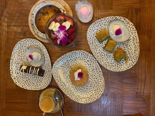 A variety of desserts on decorative plates, including cakes, fruit, and a coffee cup, arranged on a wooden table.