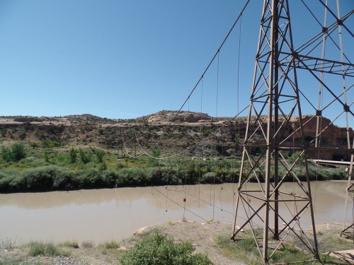 A power line tower stands near a muddy river, with rocky hills and greenery in the background under a clear blue sky.