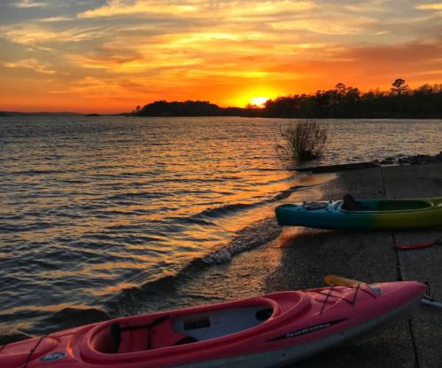 Two kayaks on a shore at sunset, with vibrant orange and yellow hues reflecting on the water.