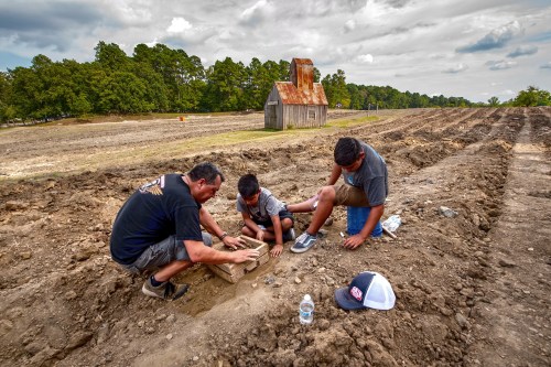 Three people, including a child, work together in a field, examining a box in the soil near a rustic barn.