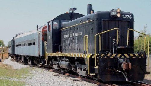 A black and yellow vintage train engine with a blue passenger car on a gravel track under a clear sky.
