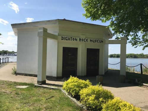 Exterior of the Dighton Rock Museum, a white building with a sign, surrounded by greenery and near a body of water.