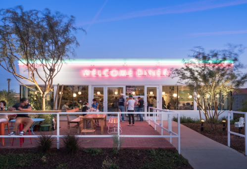 A modern diner with a neon "WELCOME DINER" sign, outdoor seating, and people enjoying the evening ambiance.
