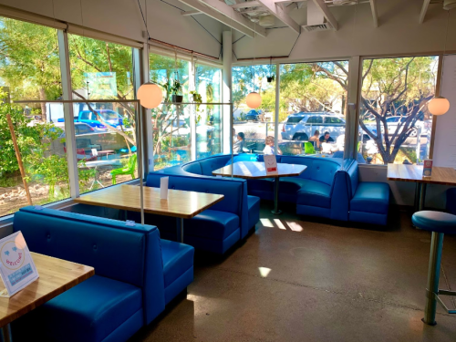 Bright, modern café interior with blue seating, wooden tables, and large windows showcasing greenery outside.