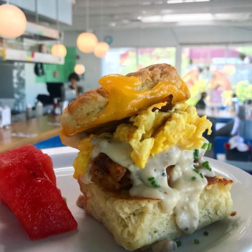 A delicious breakfast sandwich with a biscuit, scrambled eggs, cheese, and gravy, served with a slice of watermelon.