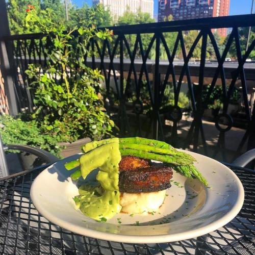 A plate of grilled chicken topped with asparagus and green sauce, served on a patio with a city view in the background.