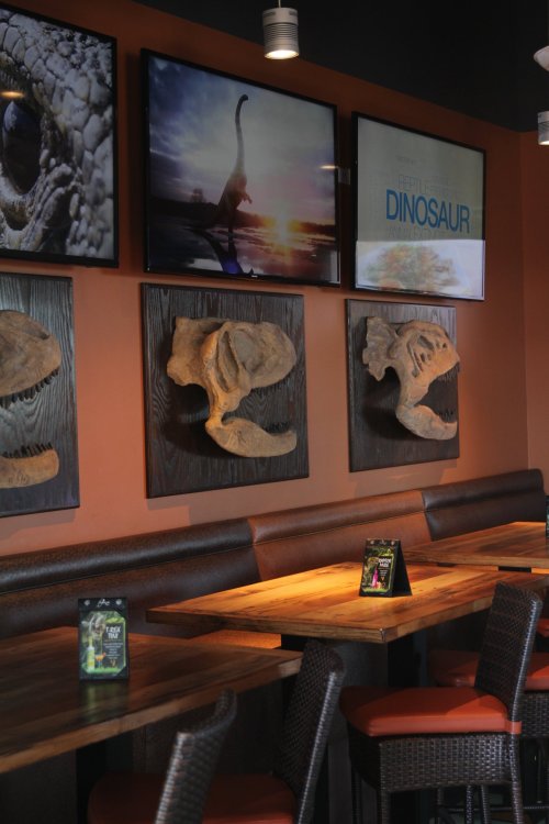 Interior of a restaurant featuring dinosaur-themed decor, including wall art and fossil replicas, with wooden tables.