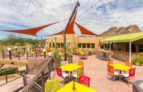 Colorful outdoor dining area with yellow and red tables, string lights, and shaded seating against a scenic backdrop.