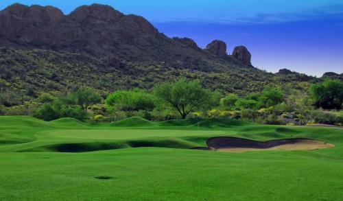 Lush green golf course with rolling hills and a sand trap, set against a backdrop of mountains and a clear blue sky.