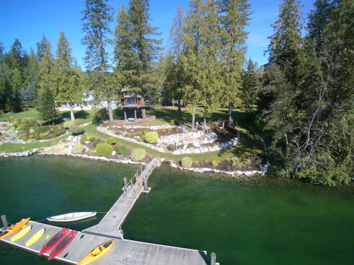 A serene lakeside view with a wooden dock, kayaks, and lush greenery surrounding a landscaped garden.