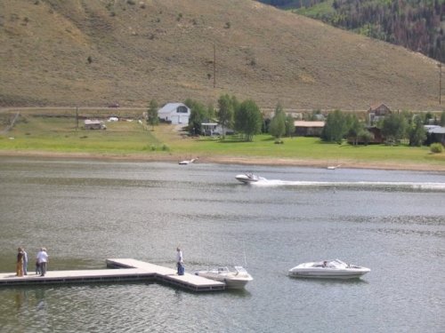 A scenic lake view with boats, a dock, and a hillside in the background, featuring green trees and a few houses.