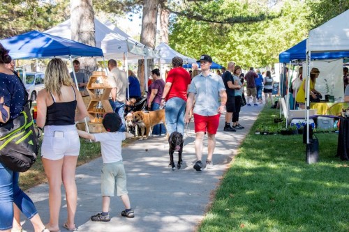 A busy outdoor market with people browsing stalls, walking dogs, and enjoying a sunny day in a park.