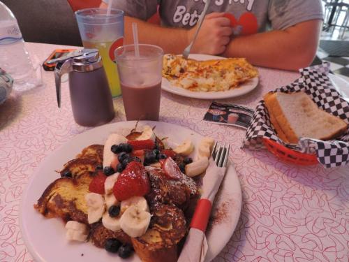 A plate of French toast topped with strawberries, bananas, and blueberries, with drinks and an omelet in the background.