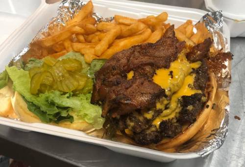 A loaded cheeseburger with bacon, lettuce, and pickles, served with crispy fries in a takeout container.