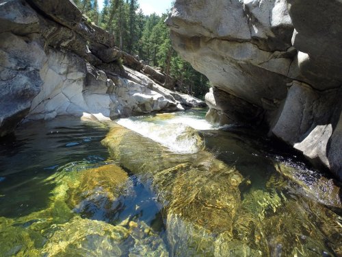 A serene river flows between rocky cliffs, surrounded by lush green trees under a clear blue sky.