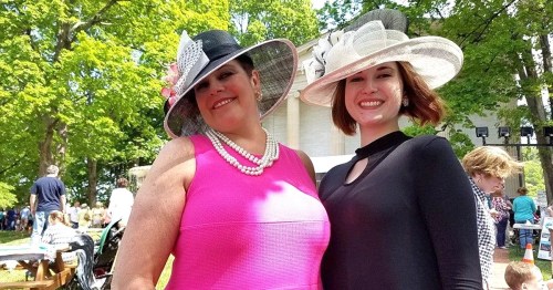 Two women in stylish hats and dresses smile at the camera, surrounded by greenery and a crowd in the background.