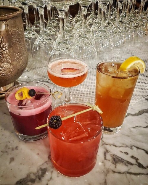 Three colorful cocktails on a marble bar, with a backdrop of sparkling glassware.