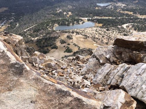 A rocky cliff edge overlooking a valley with lakes and scattered trees in a mountainous landscape.