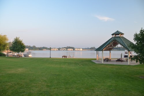 A serene lakeside view featuring a gazebo, green lawn, and boats in the distance under a clear sky.