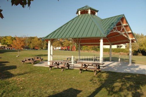 A green-roofed gazebo in a park, surrounded by picnic tables and grassy areas, with trees in the background.