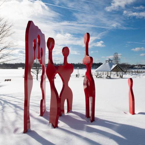 Red abstract sculptures stand in a snowy landscape, with a gazebo and trees in the background under a clear blue sky.