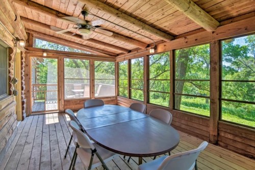 A spacious screened porch with a wooden ceiling, featuring a round table and chairs, surrounded by greenery.