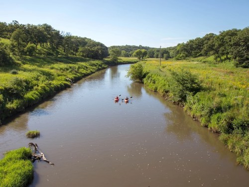 A serene river flows through lush greenery, with kayakers enjoying the calm water under a clear blue sky.