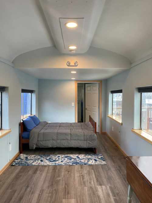 A cozy bedroom with a bed, blue bedding, large windows, and wooden flooring, featuring a simple, modern design.
