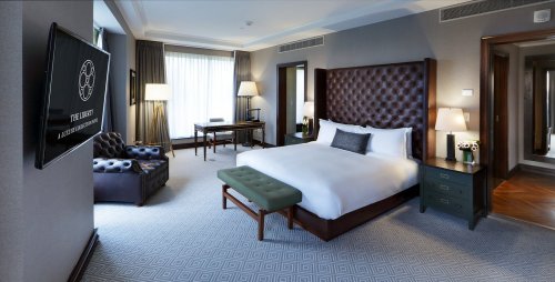 A modern hotel room featuring a king-sized bed, armchair, desk, and large windows with natural light.