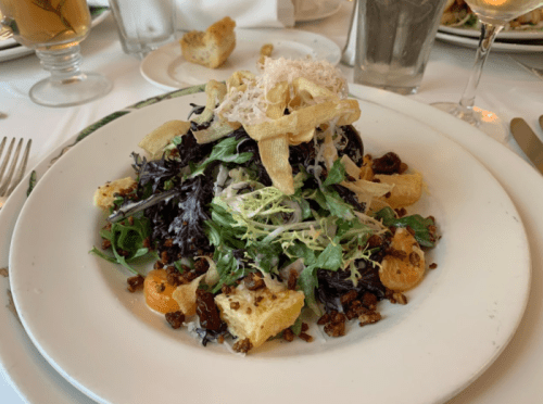 A colorful salad with mixed greens, crispy toppings, nuts, and citrus served on a white plate.