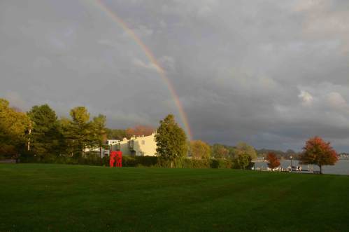 A vibrant rainbow arcs over a grassy field with trees and a building in the background, under a cloudy sky.