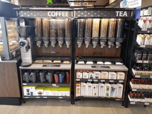 Self-serve coffee and tea station with various beans and leaves in dispensers, cups, and brewing equipment below.
