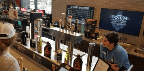 A barista serves craft beer at a counter with various taps and bottles, with a digital menu displayed in the background.