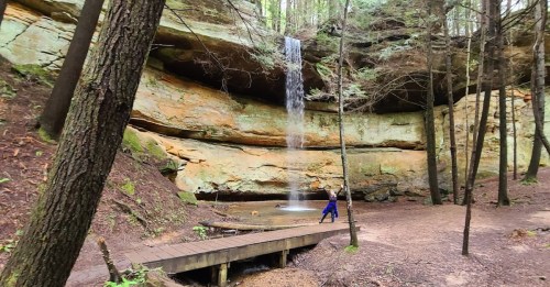 A serene waterfall cascades over a rocky cliff in a forest, with a person walking near a wooden bridge.