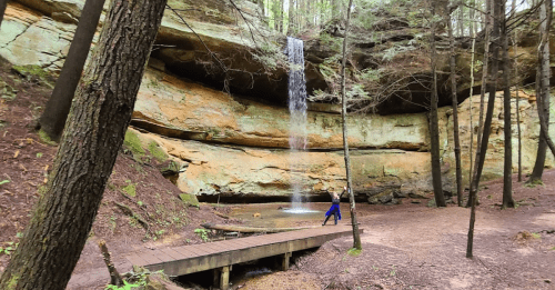 A serene waterfall cascades over a rocky cliff in a forest, with a person walking near a wooden bridge.