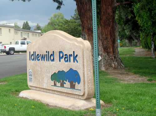 Sign for Idlewild Park with trees and grass in the foreground, set against a backdrop of buildings and trees.